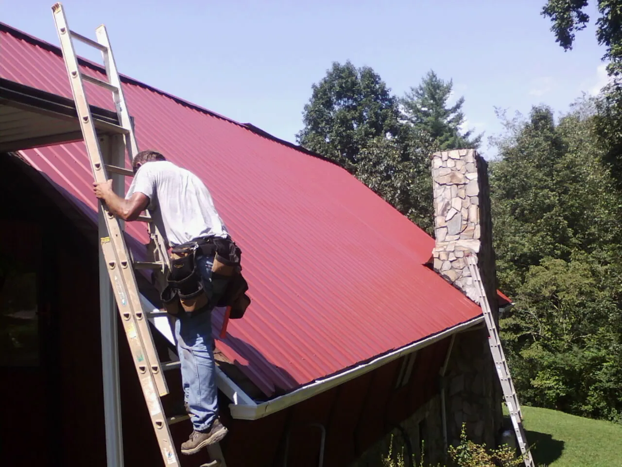 Roofer on a ladder beside a red metal roof and stone chimney — Soffit & Fascia Repair by Roofing Brothers in Long Pond